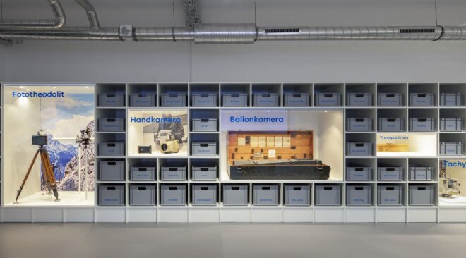 Regalwand mit grauen Boxen unterbrochen durch Bilder Theodolit, Handkamera, Ballonkamera, Transportkiste